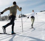 Two people cross-country skiing on a snow-covered track with trees in the background.
